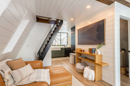 Cozy living space with a brown leather sofa, wooden shelves, and a staircase leading to an upper level. Bright and inviting.